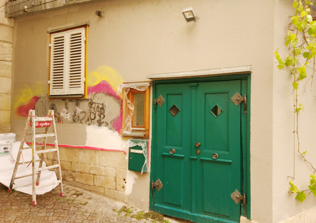 ein Graffiti auf einer Fassade mit grüner Tür ist bereits teilweise entfernt